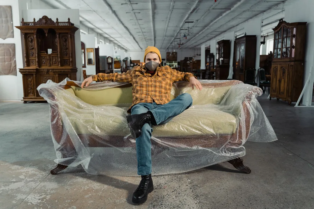 Mann sitzt auf einem in Luftpolsterfolie gepackten Sofa in einer Lagerhalle voller Schränke