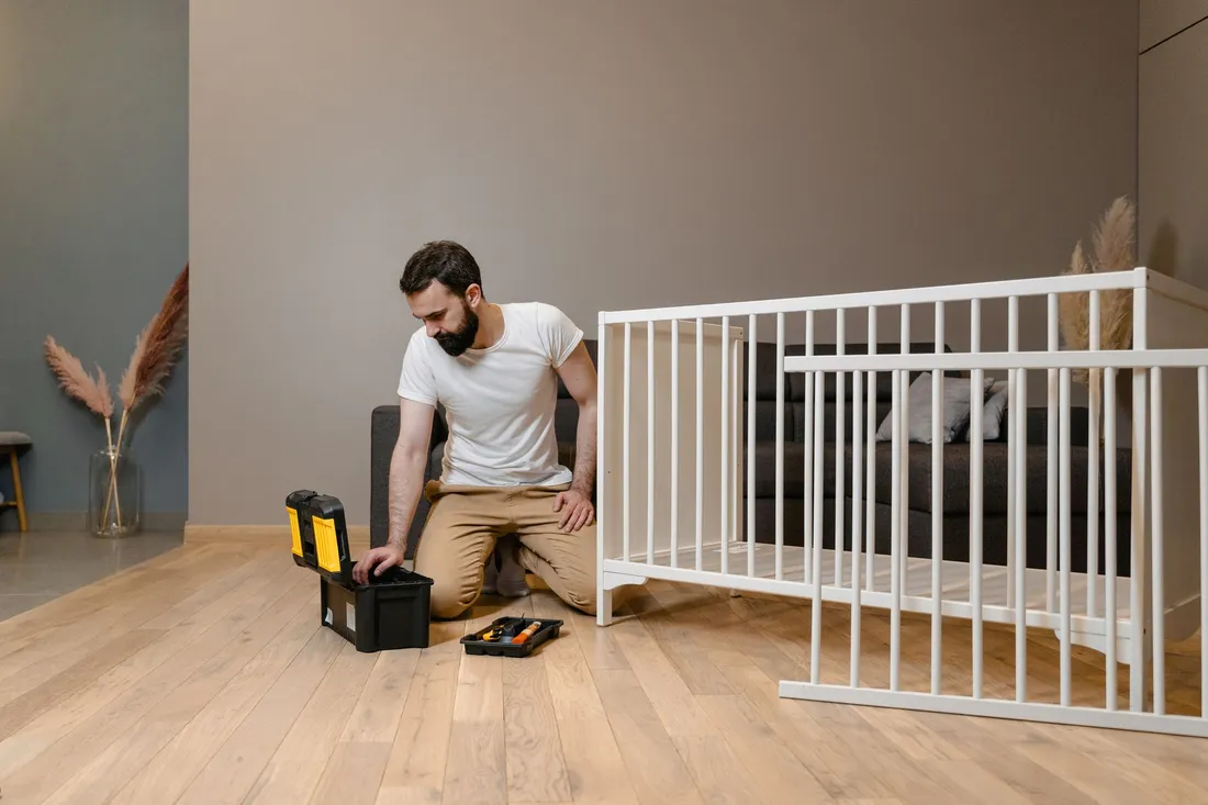 Mann kniet mit Werkzeugkoffer vor einem halb aufgebauten weißen Gitterbett