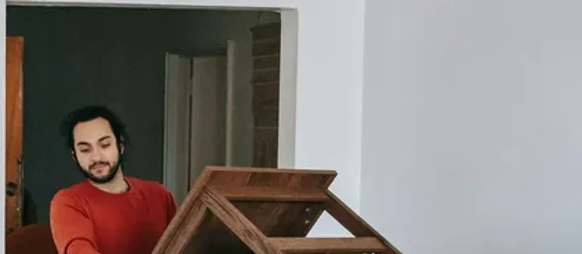 Mann im roten Pullover hält ein gekipptes Holzmöbel vor einer dunklen Wand