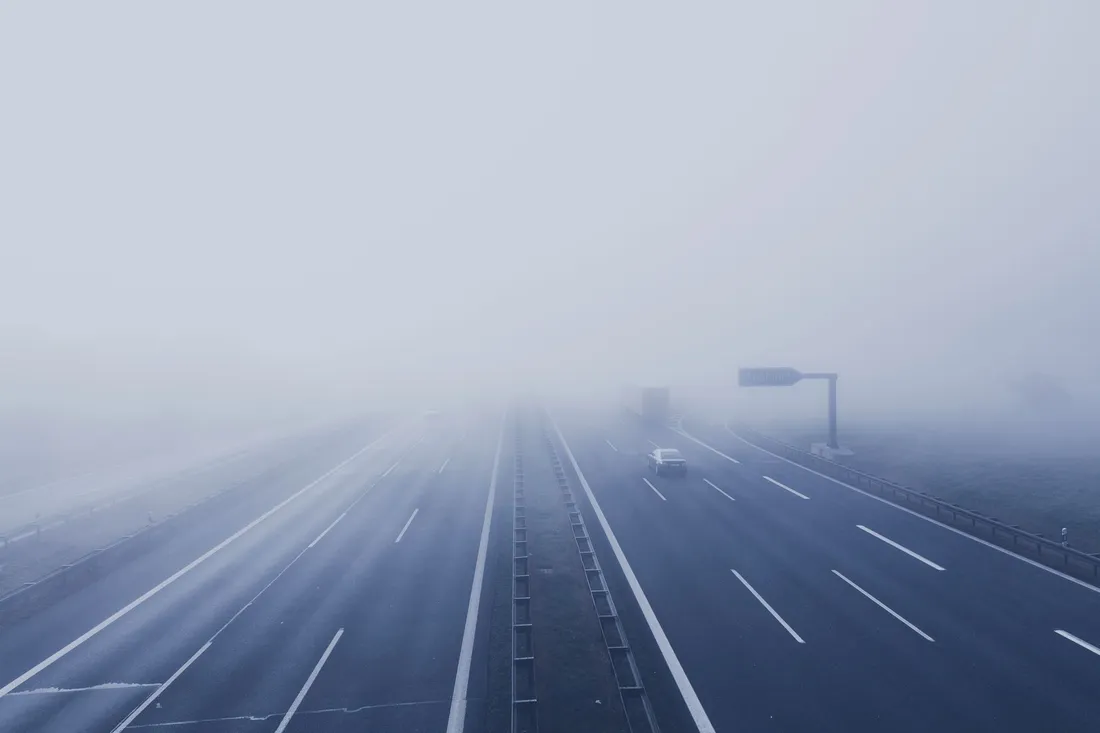 Graue Autobahn mit Mittelleitplanke, ein Lkw löst sich weit vorn aus dem Nebel
