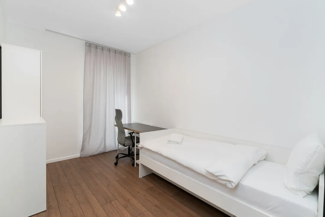 Kleines Zimmer mit Einzelbett, Schreibtisch und Drehstuhl auf Holzdielen